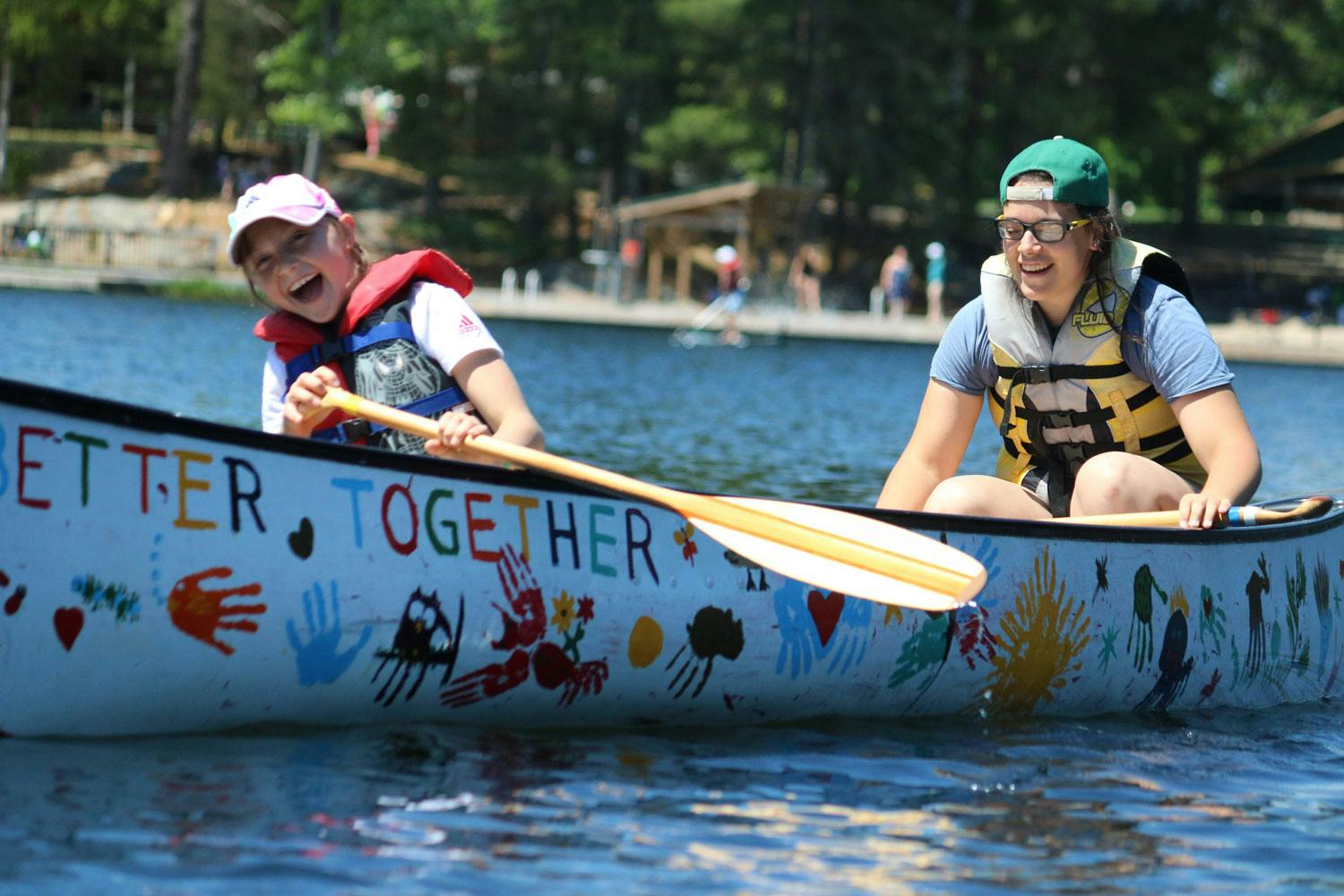 A child and a camp counsellor row a canoe together. A forest is visible in the background.