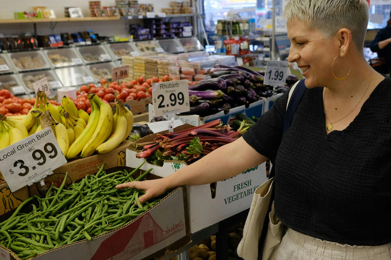 Emily Sproule puts her hand into a box of green beans in a grocery store, near boxes of rhubarb, eggplant, bananas, tomatoes.