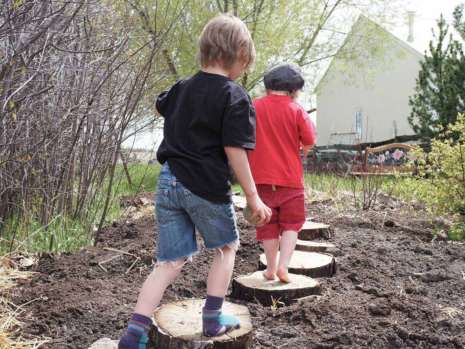 2 kids walk along a path made of log rounds between bushes in their backyard. In the distance is a bench and a white house.