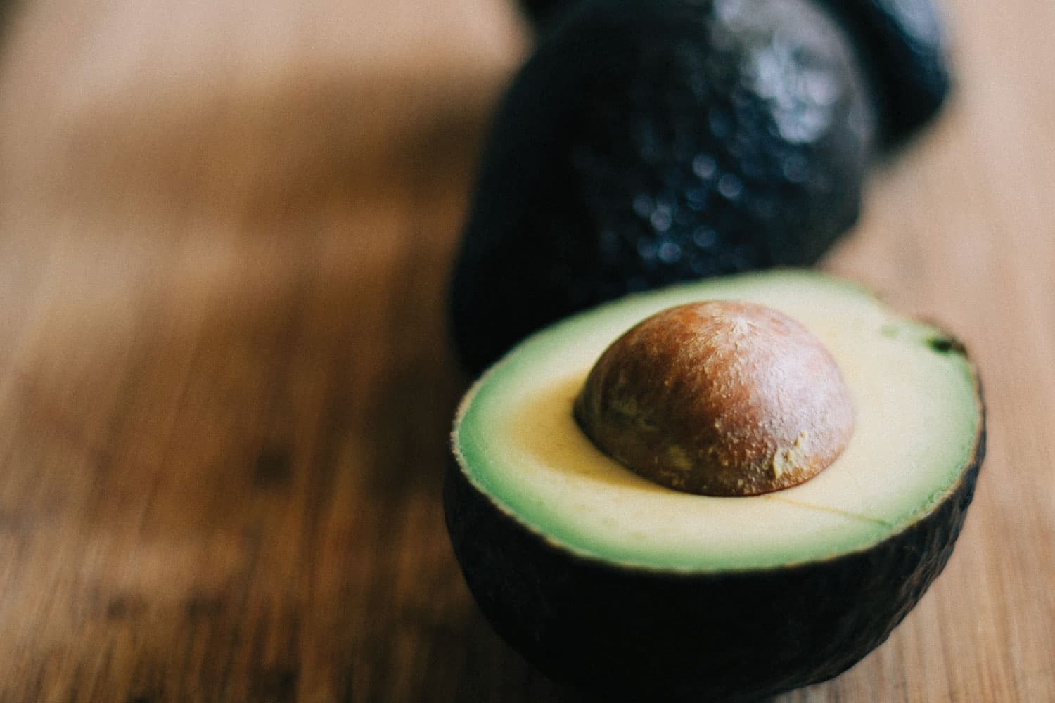 3 avocados on a wooden table. In the foreground, a cut avocado half shows light yellow-green flesh and a round, brown pit.