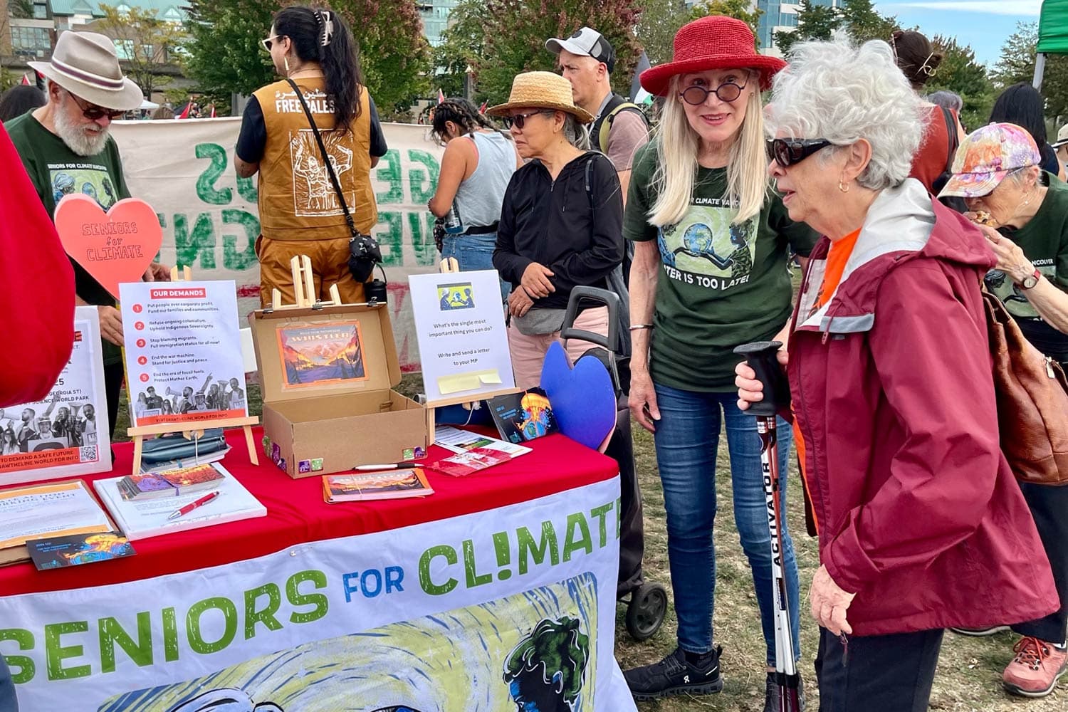 A group of older adults participate in a Seniors for Climate event outdoors. Some participants stand near a red information table displaying climate action materials, signs, and pamphlets, while others gather nearby. Trees, grass, and city buildings are visible in the background.