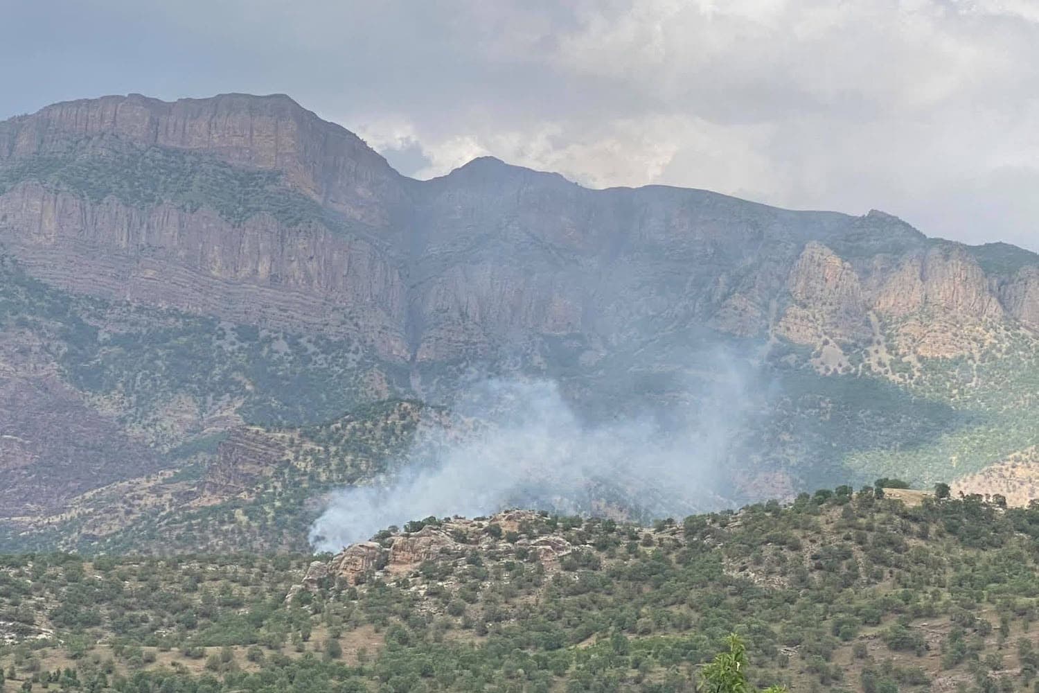 Smoke rises from a fire in a green mountain valley surrounded by steep, rocky cliffs.