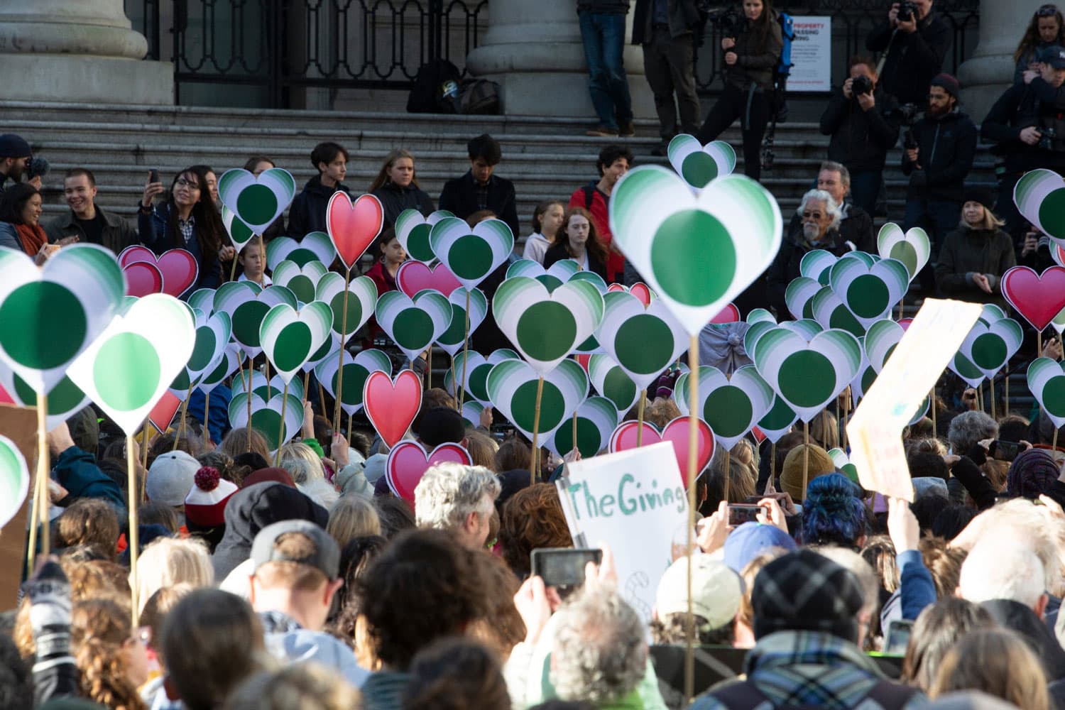 A large crowd at a climate rally holds green and red heart-shaped signs in the air, with people gathered on the steps of a building in the background.