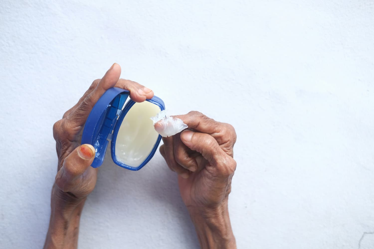 Close-up of a pair of dark-skinned, wrinkled hands holding an open blue container of petroleum jelly. One hand grips the container while the other uses an index finger to scoop out a thick, translucent glob of jelly. The hands rest against a softly textured white background.
