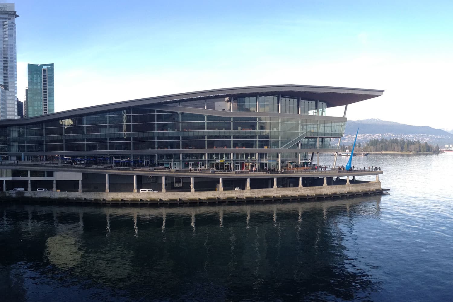 A wide, wedge-shaped four-storey building with wrap-around glass walls sits on the south shore of Vancouver Harbour.