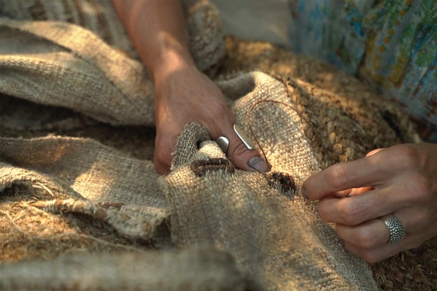 Sharon Kallis's hands sewing panels of woven nettle into a coat. She wears rings on her right thumb and left ring finger.