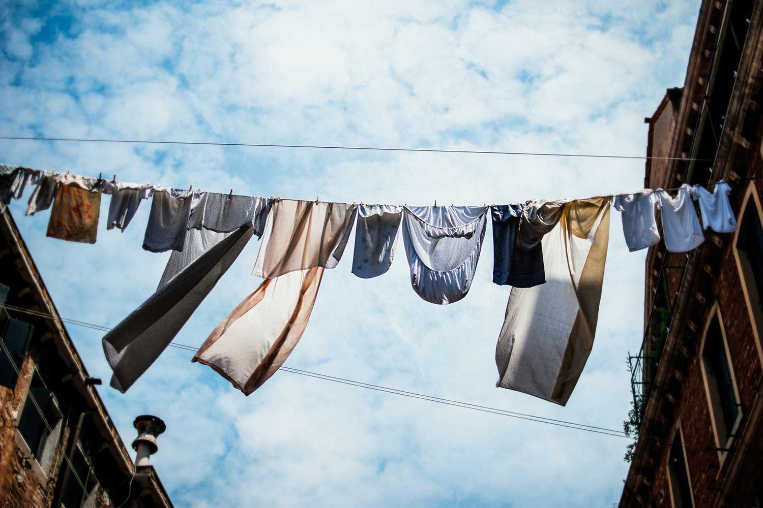 T-shirts and bed linens hang on a line stretched between two old buildings, under a bright blue sky with fluffy clouds.