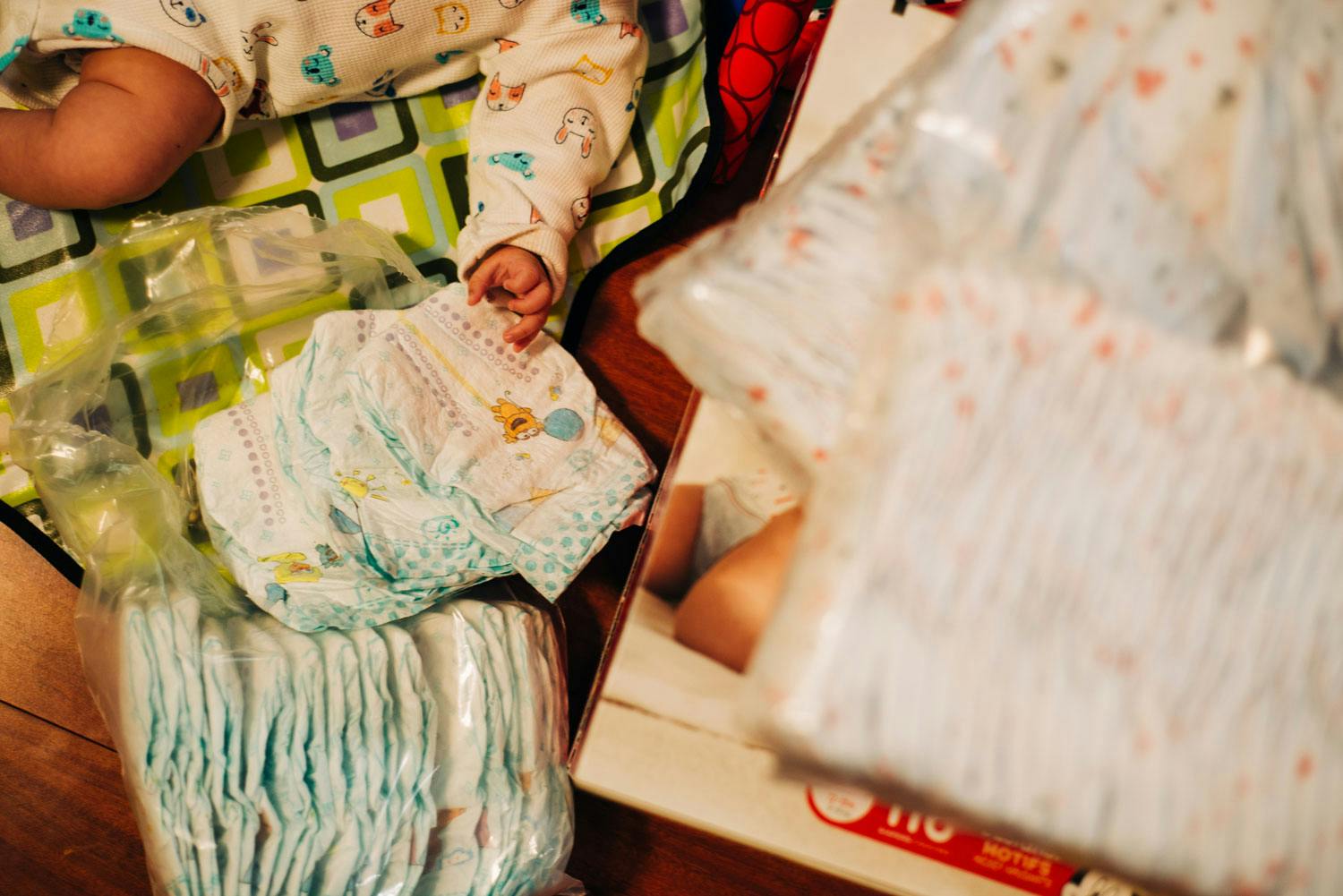 An infant wearing a onesie patterned with animal faces lies on a table beside a plastic bag of folded disposable diapers.