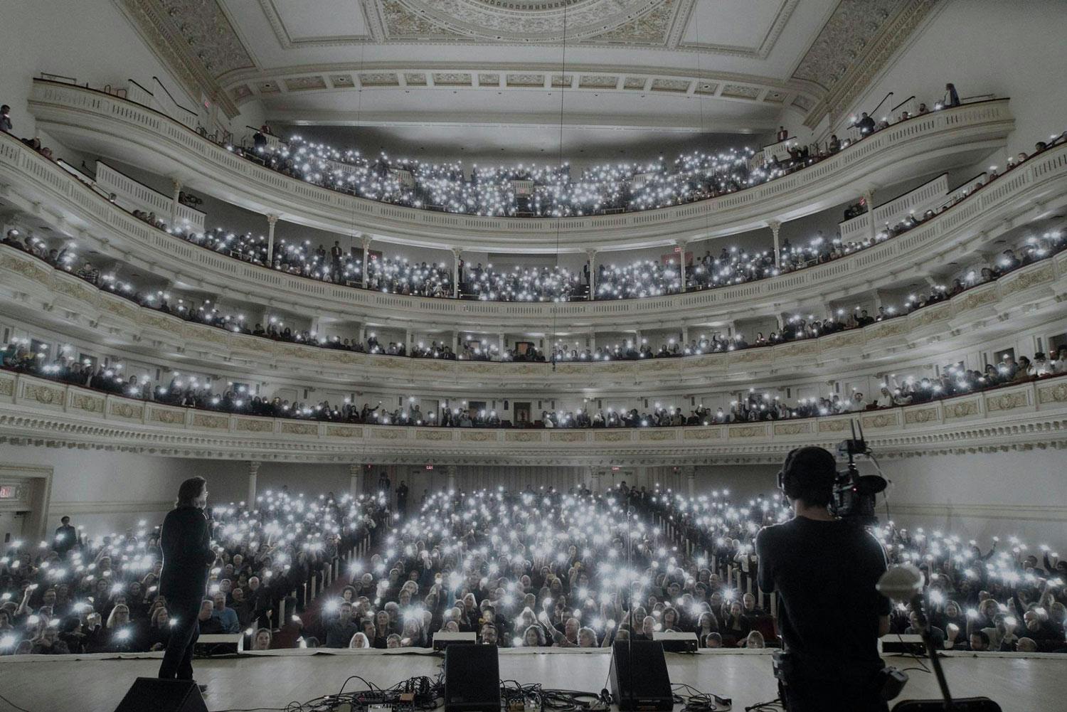 A packed, multi-storey auditorium of people hold small lamps, lighting up the entire space.