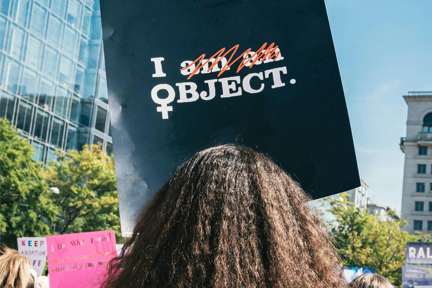 A woman with a sign bearing the text "I object" in white color seen from behind.