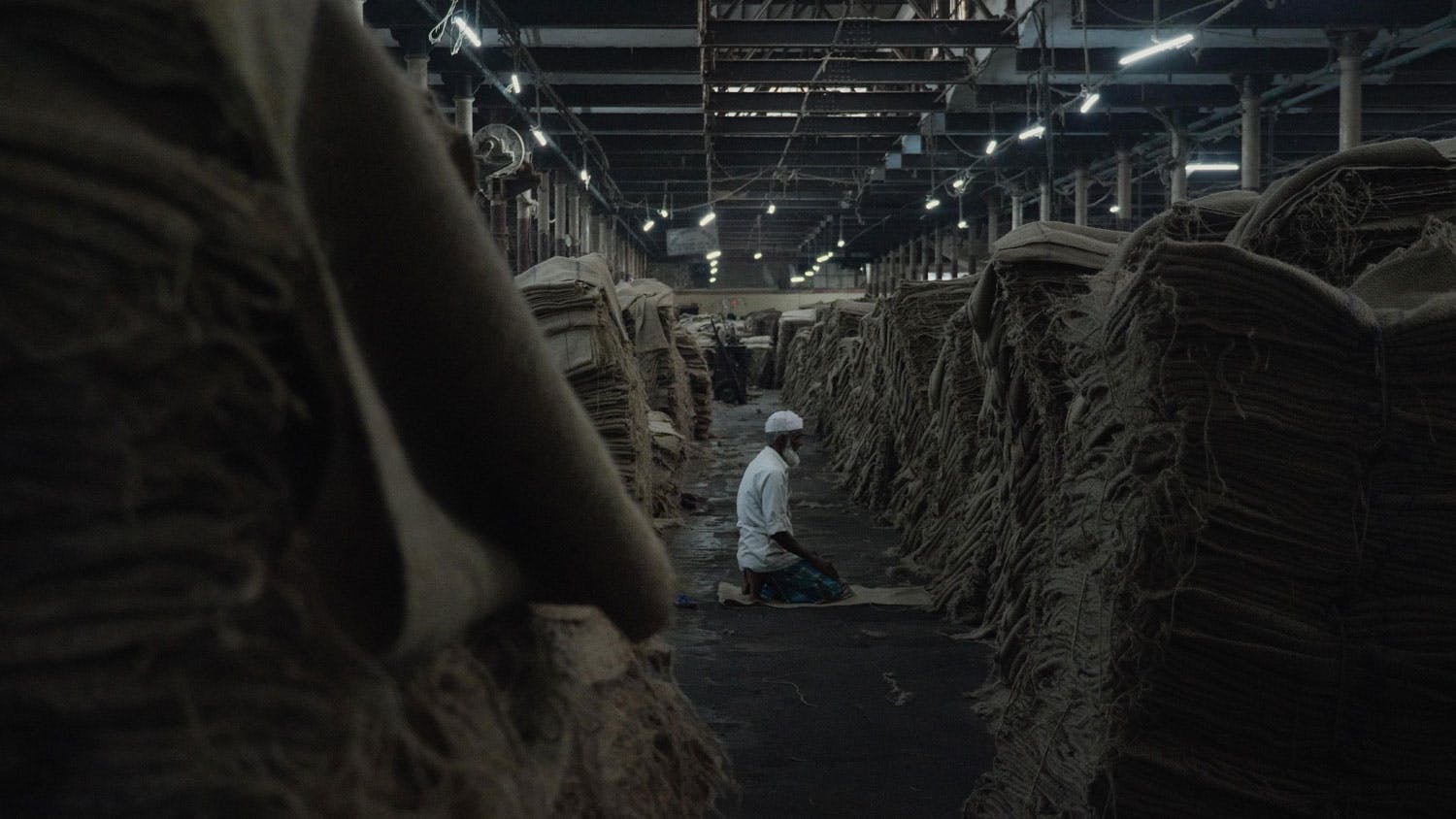 A man offers prayers on Hukumchand Mill's floor surrounded by jute stacks.