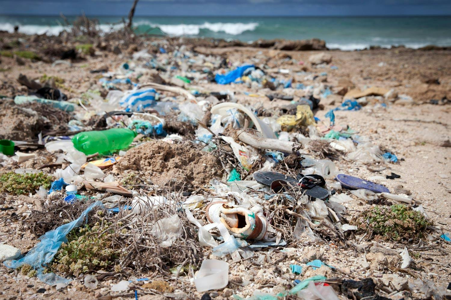 Beach covered with plastic bottles, shoes & plastic bags—blue sky and cresting turquoise ocean waves visible in background